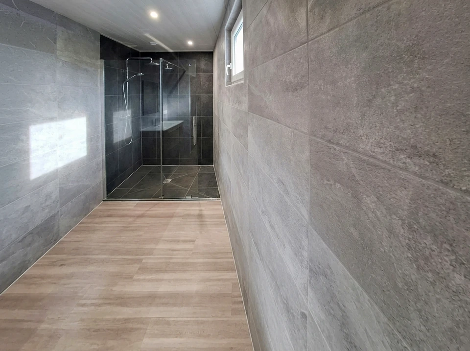 Une équipe de 2 à 3 professionnels du bâtiment travaillant ensemble dans une salle de bains en rénovation : un plombier effectuant des travaux sur les tuyauteries, un carreleur posant du carrelage mural, et un autre professionnel en arrière-plan. La scène montre clairement une coordination harmonieuse entre les différents corps de métier sur un même chantier, illustrant la maîtrise intégrale et interne de tous les domaines de compétence (plomberie, carrelage, et autres métiers du bâtiment).